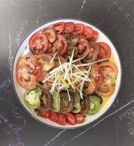 A white bowl of mixed heirloom tomato varieties &mdash; red, green and brown &mdash; on a grey marble worktop